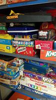 Shelf with stacked boxes of many board games including Crossfire, Scrabble Dictionary, Singlets Spirograph, Pictionary Junior, Knockout, Yahtzee, Dominoes.