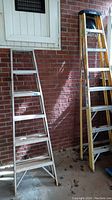 Two standing ladders shown side by side against a brick wall, one yellow fiberglass 8-foot and one metallic silver 6-foot ladder.