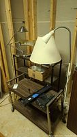 Photo showing a metal shelving unit with three shelves on wheels, with multiple lamps and lamp shades placed on and around it in a basement with unfinished walls.