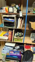 Shelves filled with office supplies including binders, folders, books, papers, and containers with small items like pencils and scissors.