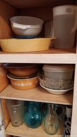 Wood kitchen cabinet shelves with white and yellow Pyrex dishes, plastic strainer, and large white plastic pitcher