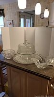 Photo of glass cake plate with dome, clear glass serving tray, clear ribbed glass bowl, and two clear plastic candy dishes on kitchen countertop with white backdrop.