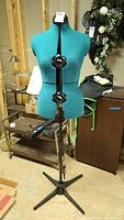 Full front view of turquoise adjustable sewing mannequin on metal tripod stand in basement setting.