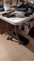 Side view of white Lifetime folding table with tools and boxes on top and tripod under it.