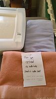 Photo showing pink twin size wool blanket with satin trim folded with note describing fabric and condition, next to lavender cotton blanket and plastic box lid