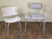 Two adjustable bath/shower chairs: one white plastic with holes and backrest, one grey padded with backrest and side rail, both on adjustable legs with rubber tips.