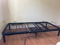 Black metal twin size adjustable bed frame with mesh base, wired remote control and power cord visible, placed on wooden floor against beige wall.