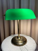 Front view of bankers lamp with green glass shade and brass base on round marble table.