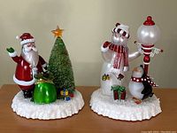 Wide view displaying both Christmas lighted figurines on snow-textured bases: Santa Claus with tree and gifts on left, snowman with penguin and lamp post on right.