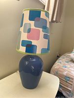 Blue ceramic lamp with a colorful geometric pattern lampshade showing squares and rectangles in blue, pink, and green, placed on a small table.