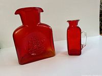 Photo showing large cranberry glass carafe with sailboat design and smaller matching cranberry mini pitcher with clear handle.