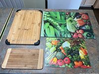 Five cutting boards displayed on a white surface: three glass vegetable print boards and two wooden boards with different sizes and corner grips.