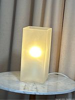 Lit rectangular frosted glass lamp on a marble surface with curtains in the background.