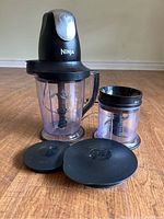 Overhead view of two clear blending containers with black handles, blade assemblies inside, and two lids on floor in front. One container is larger at 6-cups, the other smaller at 2-cups, alongside a black motor base.