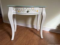 Front view showing white painted table with curved legs, floral vine hand painted designs in green and yellow on drawer front and around top edge, front drawer closed with green knob.