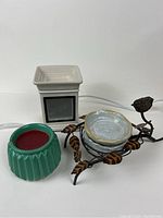 Two ceramic Scentsy warmers, white square with ventilation holes and teal ridged warmer. Decorative metal stand with bird and leaves holding four small ceramic plates.