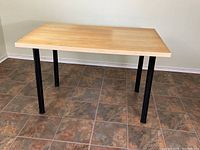 Full side view of rectangular table with light wood veneer top and four black metal legs on tiled floor.