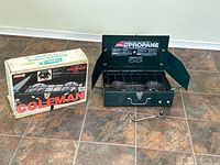 Photo of the Coleman Tourist 2 burner propane stove with original box to the left, placed on tiled floor.