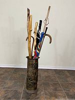 Photo shows vintage metal umbrella stand filled with walking sticks and umbrellas, highlighting embossed design on stand.