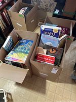 Three cardboard boxes filled with assorted books, top covers visible including Van Gogh art book, Mayo Clinic, 101 Catastrophes, Toronto guide, and other titles.