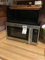Hamilton Beach stainless steel microwave and black metal bread box on wooden countertop near brick wall.