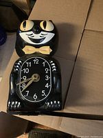 Front view showing the black cat clock with yellow eyes and bow tie, clock face is visible with white numbers.