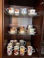 Three shelves inside a wooden cabinet holding assorted china teacups and porcelain mugs with floral designs. Includes some Royal Albert mugs and orphan cups.