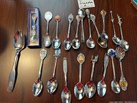 Top-down view of 18 collector souvenir spoons in stainless steel and silver plate arranged on wood surface including one boxed spoon and various enamel and sculpted handles.