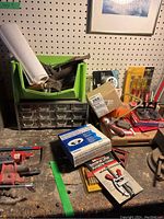 Wide view showing green plastic bin with various hand tools, red metal toolbox, hardware drawers, screwdrivers, and terminal lifter box on a workbench under a pegboard wall.