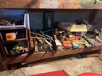 Full view of wooden workbench lower shelf filled with assorted hand tools, fasteners, and hardware items including nails, screws, wrenches, and clamps.