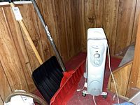 Image showing the heater and two snow shovels against wood-panel walls with carpeted floor. Heater is white with visible controls and indicator light. Two shovels include a black plastic blade shovel with wooden handle and a red plastic blade shovel with metal handle.