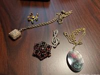 Overview of all five jewelry pieces on wooden surface showing the two necklaces, two brooches, and earrings.