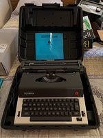 Olympia Electric typewriter housed in a hard plastic case, with power cord attached, viewed from the keyboard side.