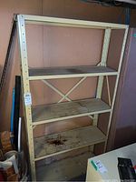 Full view of the shelving unit showing four shelves with wear and stains, beige metal frame, placed against a wall in basement.