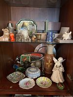Shelf with assorted Wedgwood trinket box, ceramic animal figurines, silverplated baby cup, decorative plates, vase, candles, and other small items.