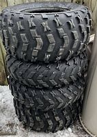 Stack of four black off-road tires with an aggressive tread pattern, placed outdoors next to a wood fence and snow on the ground. Tires show signs of use but are intact.