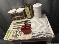 Photo of assorted kitchenware including knives, peelers, timers, bottle openers, white canister, and dishcloths on table