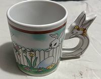 Photo showing the ceramic mug with bunny design painted on the side and a handle shaped like a bunny with a yellow ribbon.