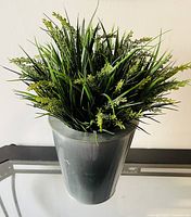 Front view of faux green plant with thin leaves and small yellow buds in gray metal planter on glass surface.