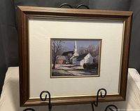 Overall view of the framed print showing the village church and buildings in daytime under a blue sky with shadows cast on the path.