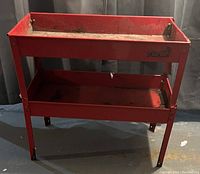 Red metal two-tier tool trays stand, showing rust and wear, against dark background.