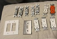 Photo of various electrical hardware items including toggle light switches, duplex outlets in white and orange colors, and switch plates in metal and white.