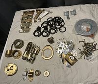 Photo showing various door hardware laid out, including hinges, deadbolt locks with keys, several metal rings, screws, and miscellaneous metal fittings on a beige surface.