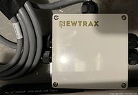 Close-up of Newtrax branded rectangular electronic module with multiple cable connectors and thick gray cable attached, placed on a gray floor.