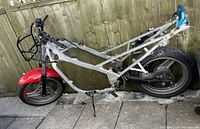 Motorbike frame missing many components, showing front and rear wheels, front red mudguard, rear blue mudguard, swingarm, and some handlebar parts