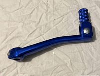 Blue anodized kick starter lever laid flat on a light fabric surface, showing knurled foot grip and attachment hole.