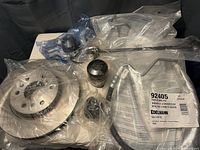 Full view of all vehicle parts including brake rotors, wheel spacers, rod components, and bushings sealed in original plastic packaging on table.