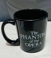 Black ceramic mug with 'The Phantom of the Opera' text in white on one side, glossy finish, rounded handle.