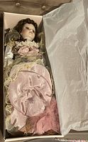 Porcelain doll wearing pink and gold vintage-style dress with lace and floral decorations, displayed lying inside original box lined with tissue paper.