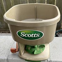 Front view of beige Scotts broadcast spreader with hopper showing brand logo, green spreader plate, and orange adjustment lever.